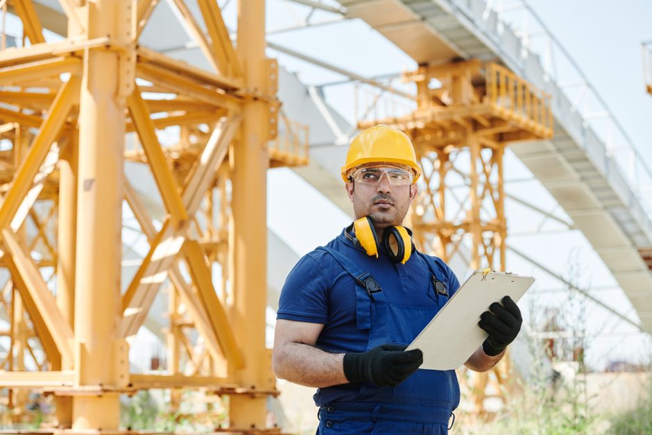 Worker in safety gear inspecting construction site with clipboard.